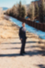 Man in a suit stands on a snowy path with hands in pockets, flanked by trees and a wooden fence under a clear sky, conveying confidence.