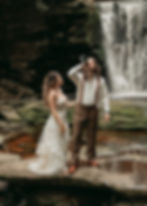 Bride in a white dress and groom in suspenders by a waterfall. Groom drinks from a bottle, bride smiles. Rocky, natural setting.