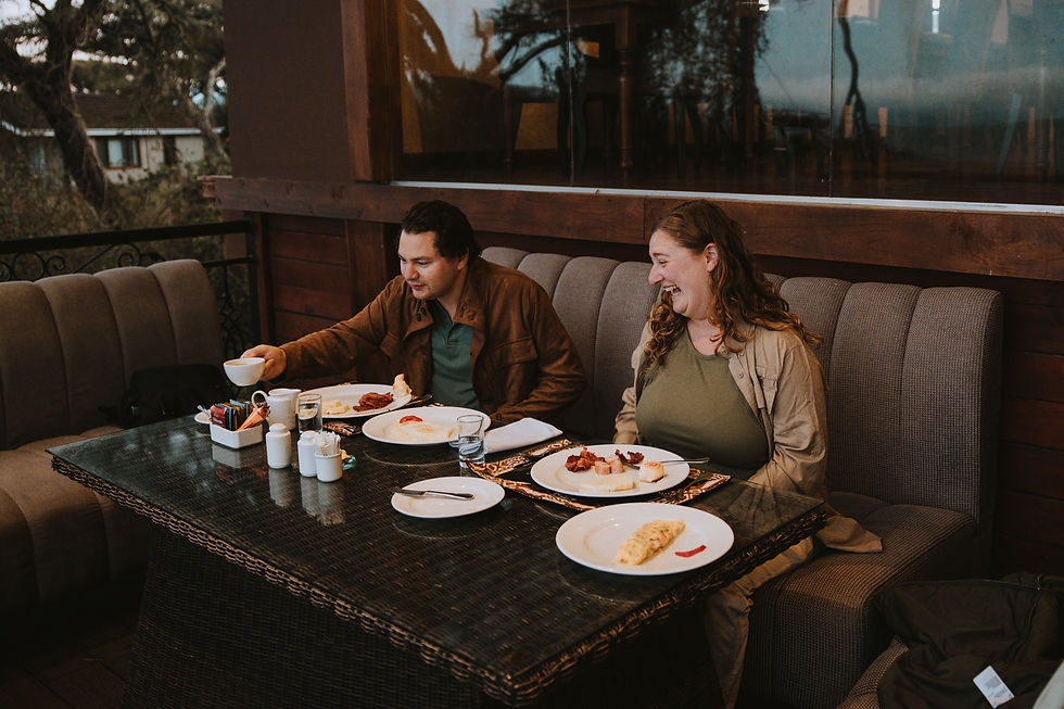 Two people dining in a cozy outdoor setting. One holds a cup, the other laughs. Plates of food and drinks on the table. Warm ambiance.
