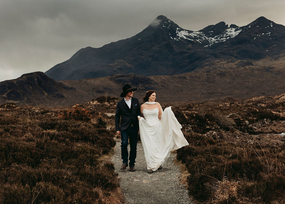 Bride and groom walk a trail in a moody landscape with the black Cuillin Mountains behind them, smiling, bride lifting her white dress, snowy peaks behind. 