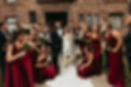 Wedding party in front of a stone building. Bride in white and groom in a tuxedo, surrounded by bridesmaids in burgundy dresses holding bouquets.