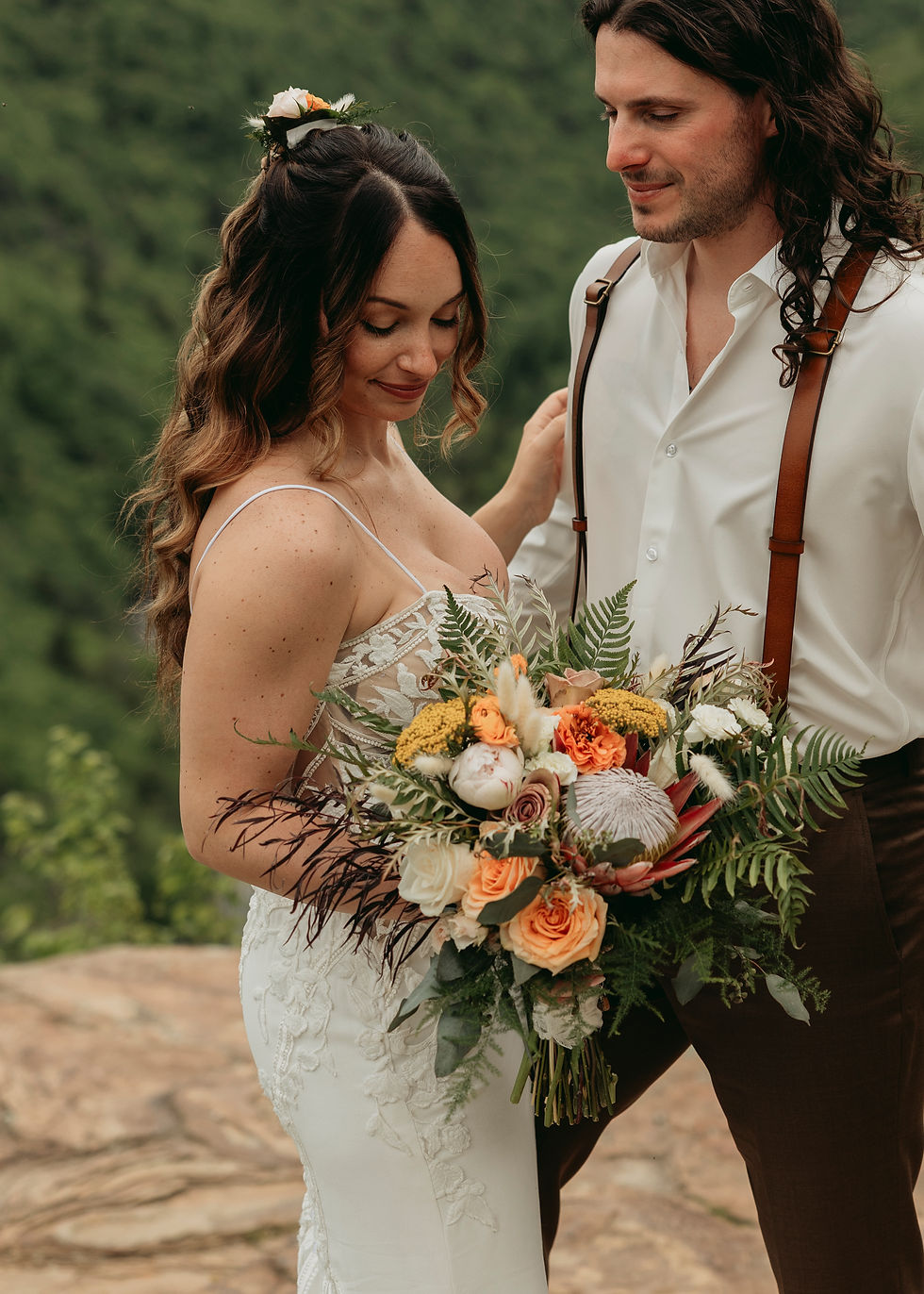 Couple embraces outdoors; the woman holds a bouquet with orange and white flowers. Both smile softly against a lush green backdrop.
