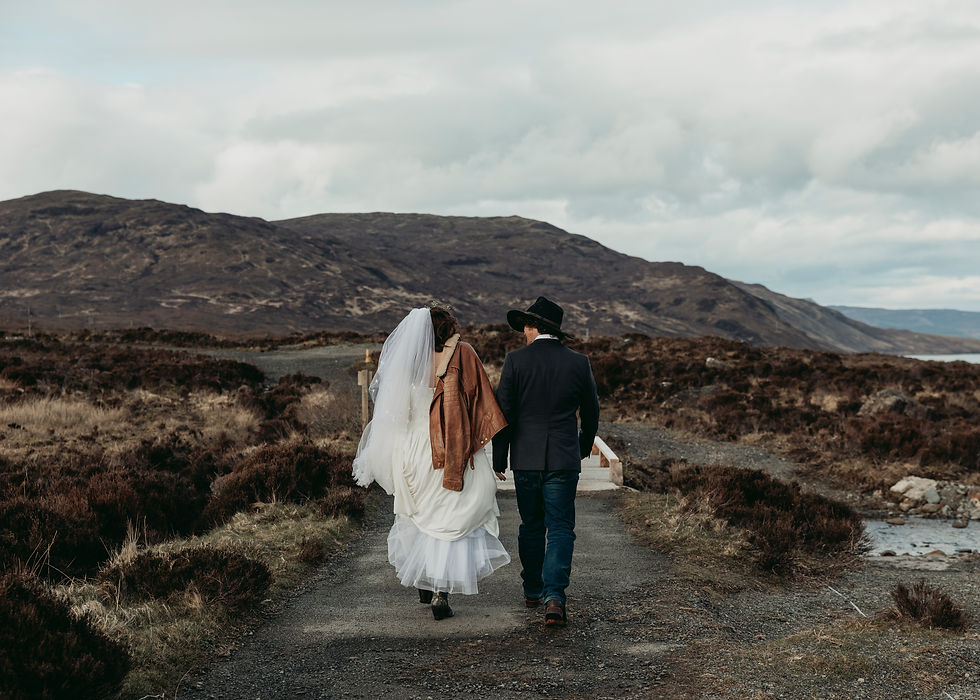Bride and groom in wedding attire walk hand in hand along a rugged mountain path under cloudy skies.