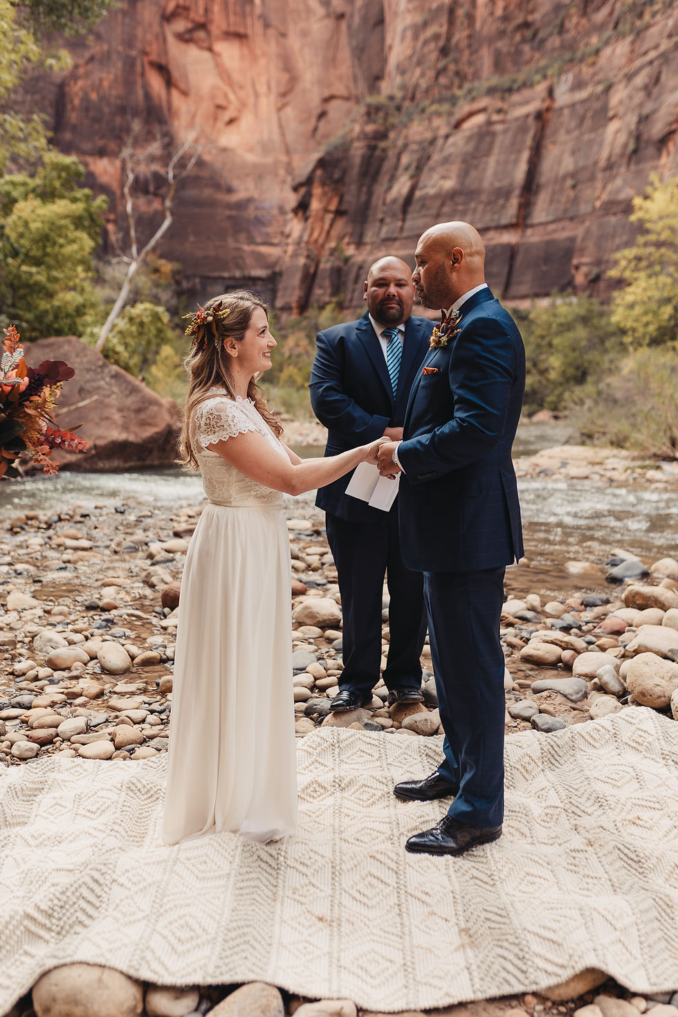 Bride and groom holding hands during a wedding ceremony by a rocky river in The Narrows in Zion National Park. An officiant stands behind. Cliffs and greenery frame the scene.