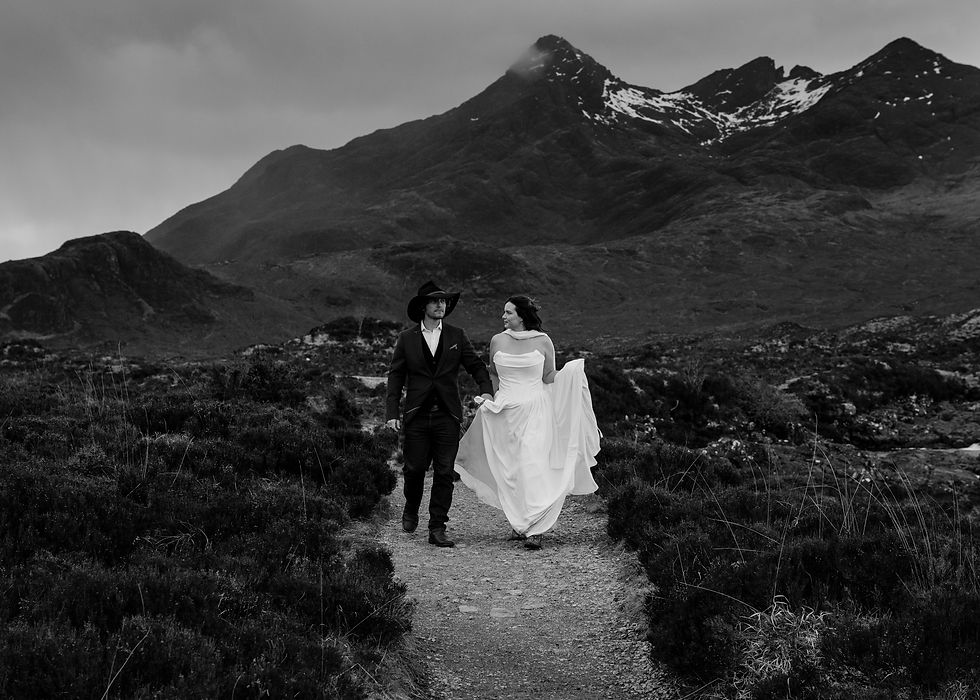 Black-and-white bride and groom walk hand in hand on a mountain path beneath snowy peaks, romantic and windswept.