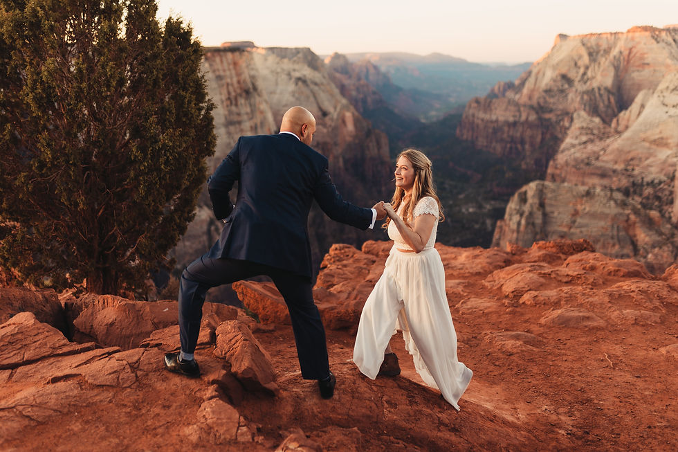 A couple on a red rock cliff in Zion National Park; man in a suit helps woman in a white dress. They smile against a canyon backdrop with a tree nearby.