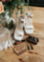 Bridal flatlay with shoes and jewelry on a wooden table, surrounded by flowers and lace.