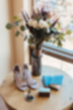 Elegant bouquet in vase by window, beige heels labeled Steve Madden, blue vow books, compact, box on wooden table; warm, serene setting.