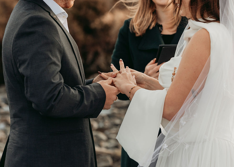 Bride and groom exchange rings during an outdoor wedding ceremony, with an officiant holding a tablet nearby.