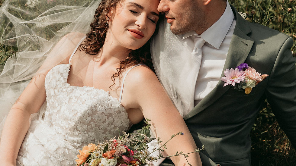 Bride in lace dress and groom in green suit lounge on grass, holding a colorful bouquet, surrounded by wildflowers, looking serene.