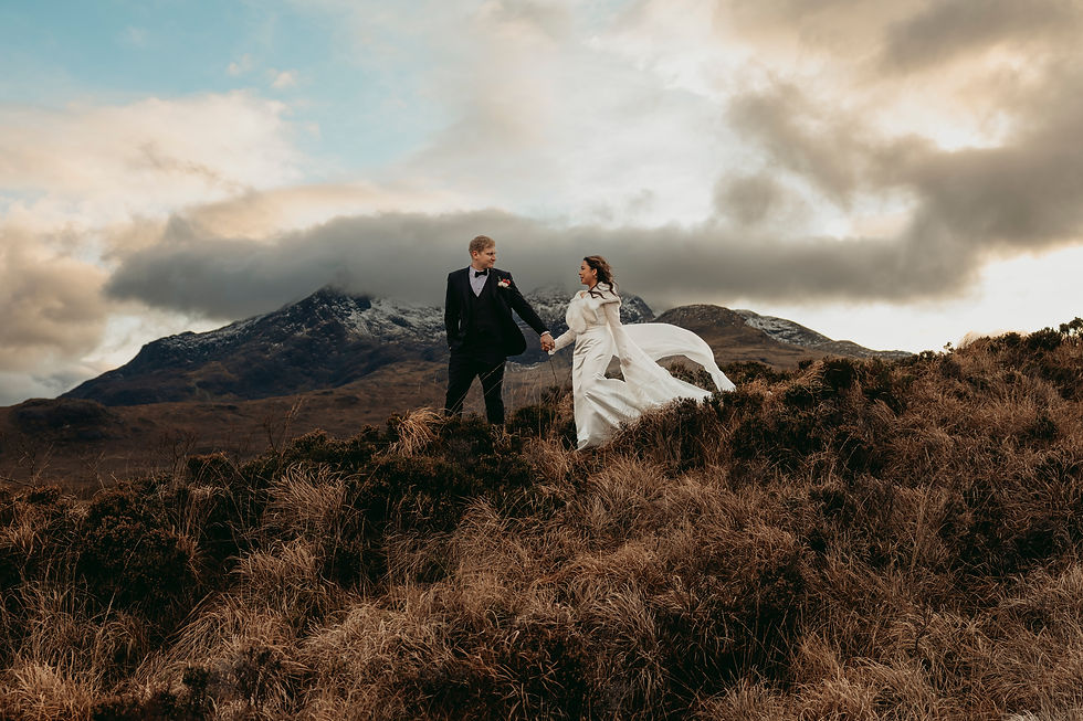 A bride and groom walking down a hill, holding hands. In the background are snowy mountains with a dramatic, cloudy sky above. 