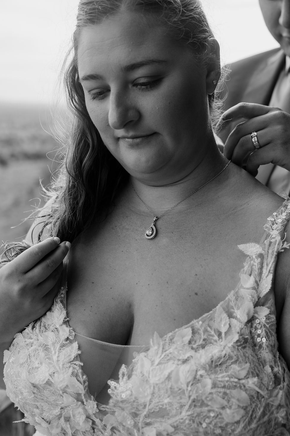 Bride in lace dress with pendant necklace, gazing down. Another person adjusts her veil. Soft background, black and white, serene mood.