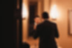 A man in a suit adjusts his boutonnière in a mirror. Warm lighting, beige walls, modern sconces, and a framed picture create a classy setting.