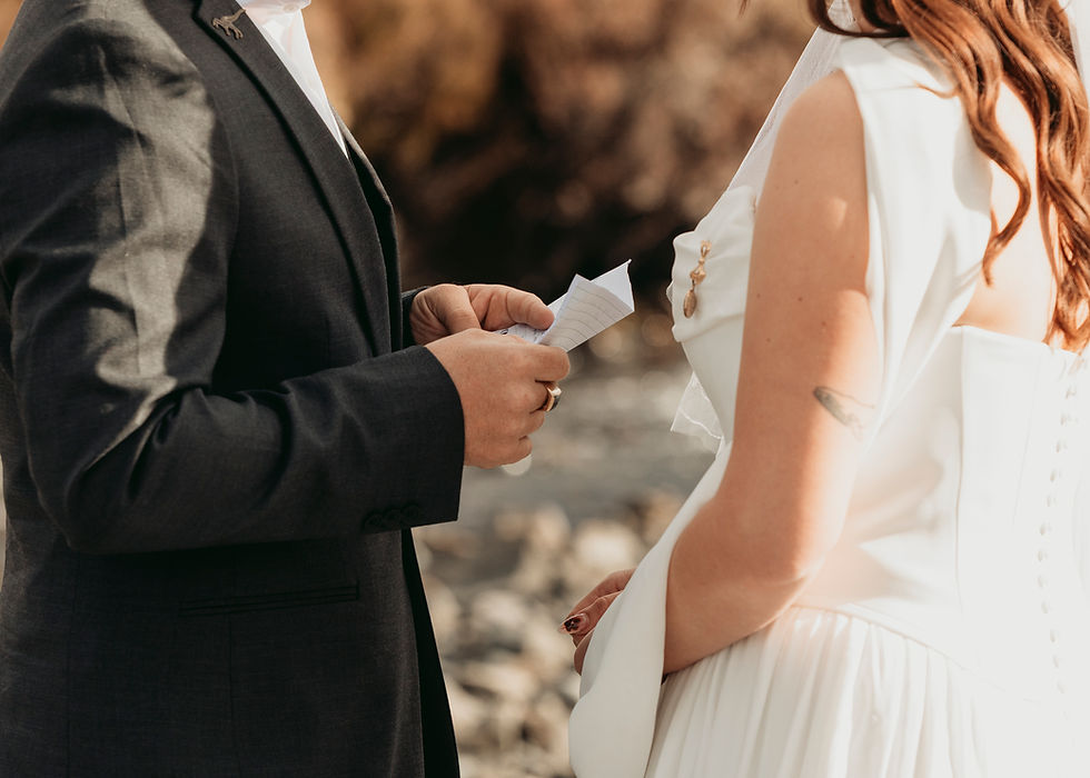 Groom in dark suit reads handwritten vows to bride in white dress outdoors, warm sunlight and blurred rocky background.