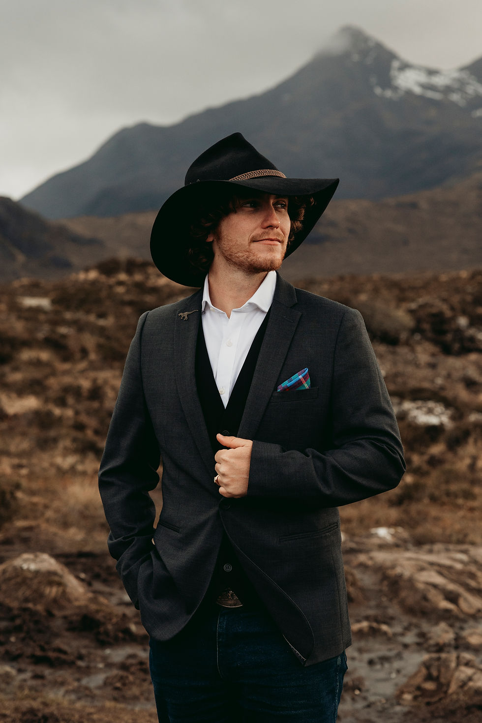 Man in a black cowboy hat and suit stands in a rocky mountain landscape, gazing off to the side with a calm, confident look.