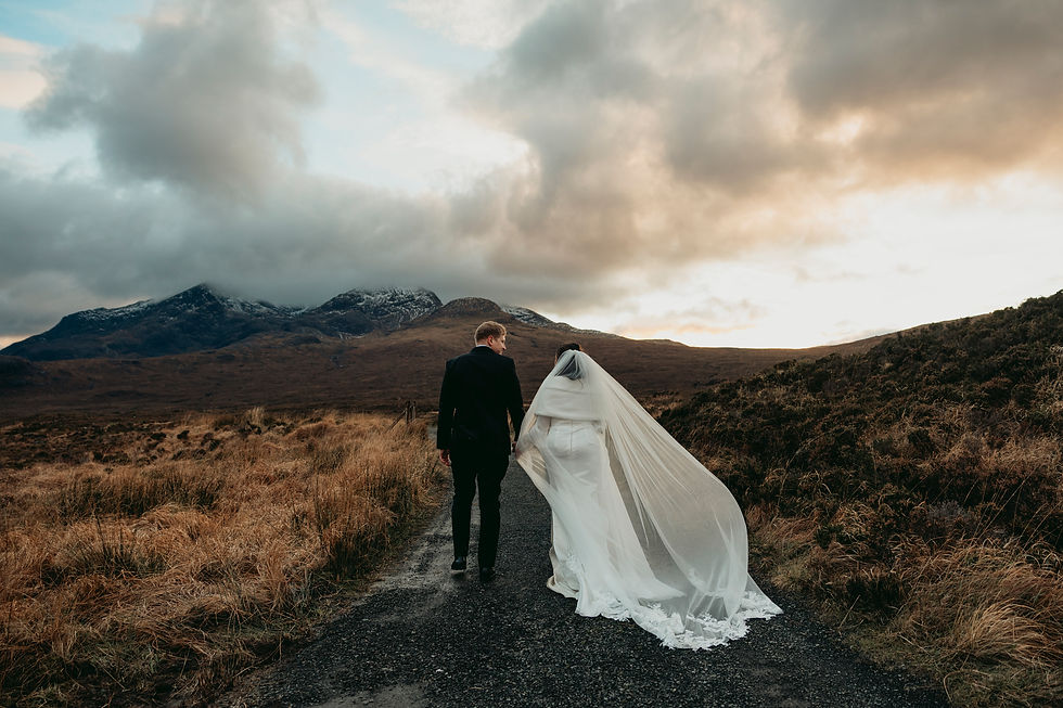 A bride and groom walk away from the camera holding hands. There are snowy mountains in the background and colorful, dramatic clouds in the sky. 
