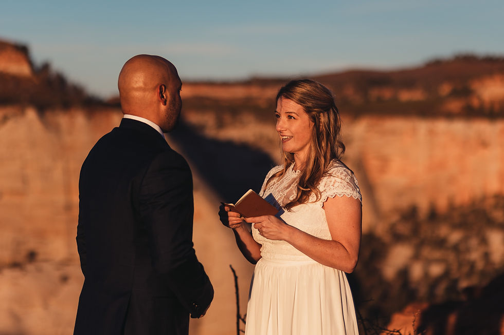 A woman in a white dress reads vows from a booklet to a man in a suit, set against a canyon backdrop at sunrise in Zion National Park, evoking a romantic mood.