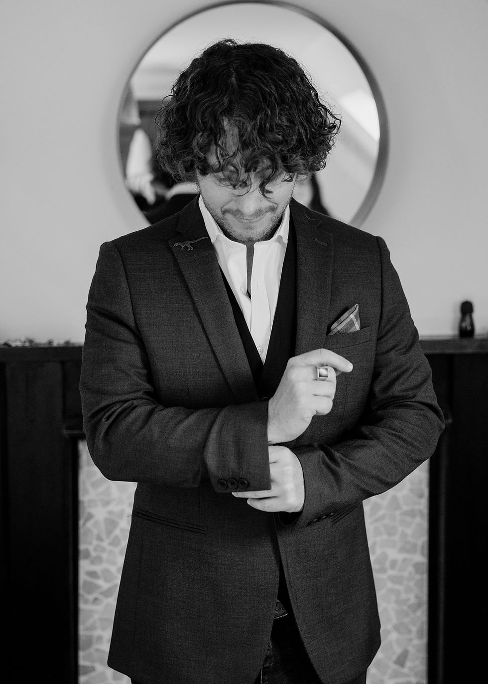 Black-and-white portrait of a groom in a suit adjusting his cuff, standing indoors before a round mirror.