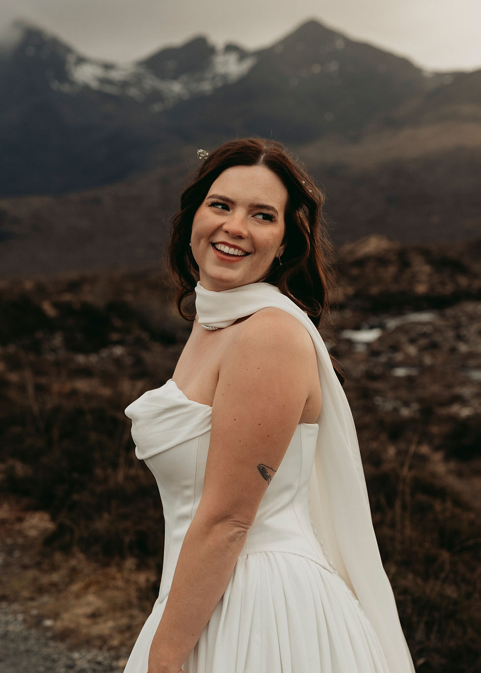 Smiling bride in a white dress and veil stands outdoors with blurred mountains behind her.