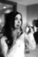 Bride applying lipstick with a brush, holding a small round mirror. She wears a lace dress. The room is softly lit, creating a serene mood.