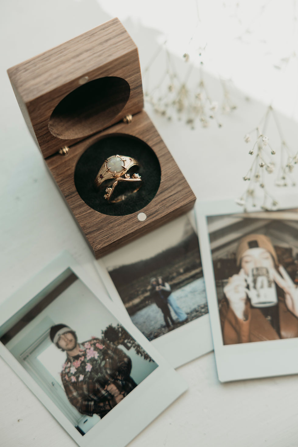 Open wooden ring box with a gold ring, surrounded by Polaroid photos and dried flowers on a white surface, soft romantic mood