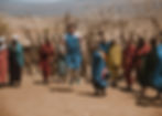 People in colorful traditional attire, smiling during a group activity outdoors. A person jumps while others watch, with a dry, rustic backdrop.