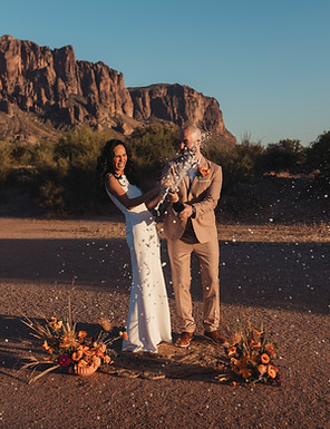 A bride and groom pop a bottle of champagne and laugh together. Behind them are the Superstition Mountains. There are flower arrangements beside them.
