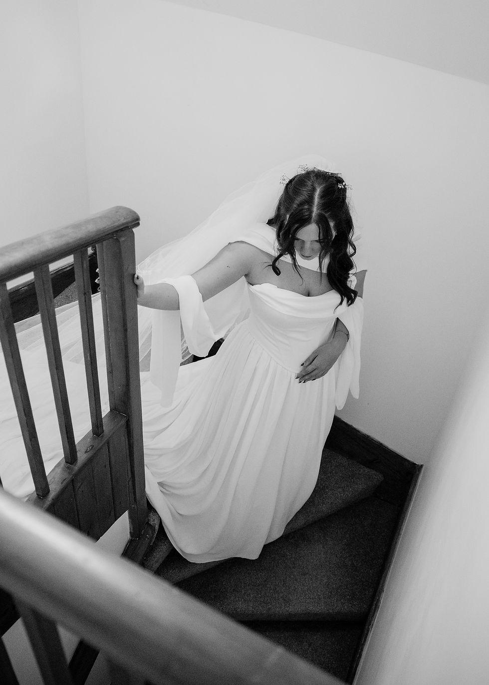 Black-and-white photo of a bride in a flowing white gown climbing a stairwell, head bowed, with a wooden banister and veil behind her.