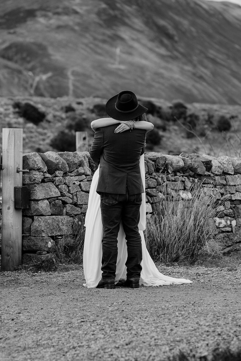 Black-and-white couple embracing by a stone wall, man in a hat and woman in a flowing dress, with mountains behind.