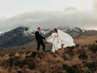 A bride and groom standing on top of a hill on the Isle of Skye in Scotland with snowcapped mountains and clouds in the background.