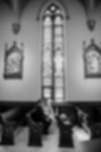 Bride and groom sit in a church pew, exchanging smiles. Stained glass windows and religious artwork in the background. Black and white tone.
