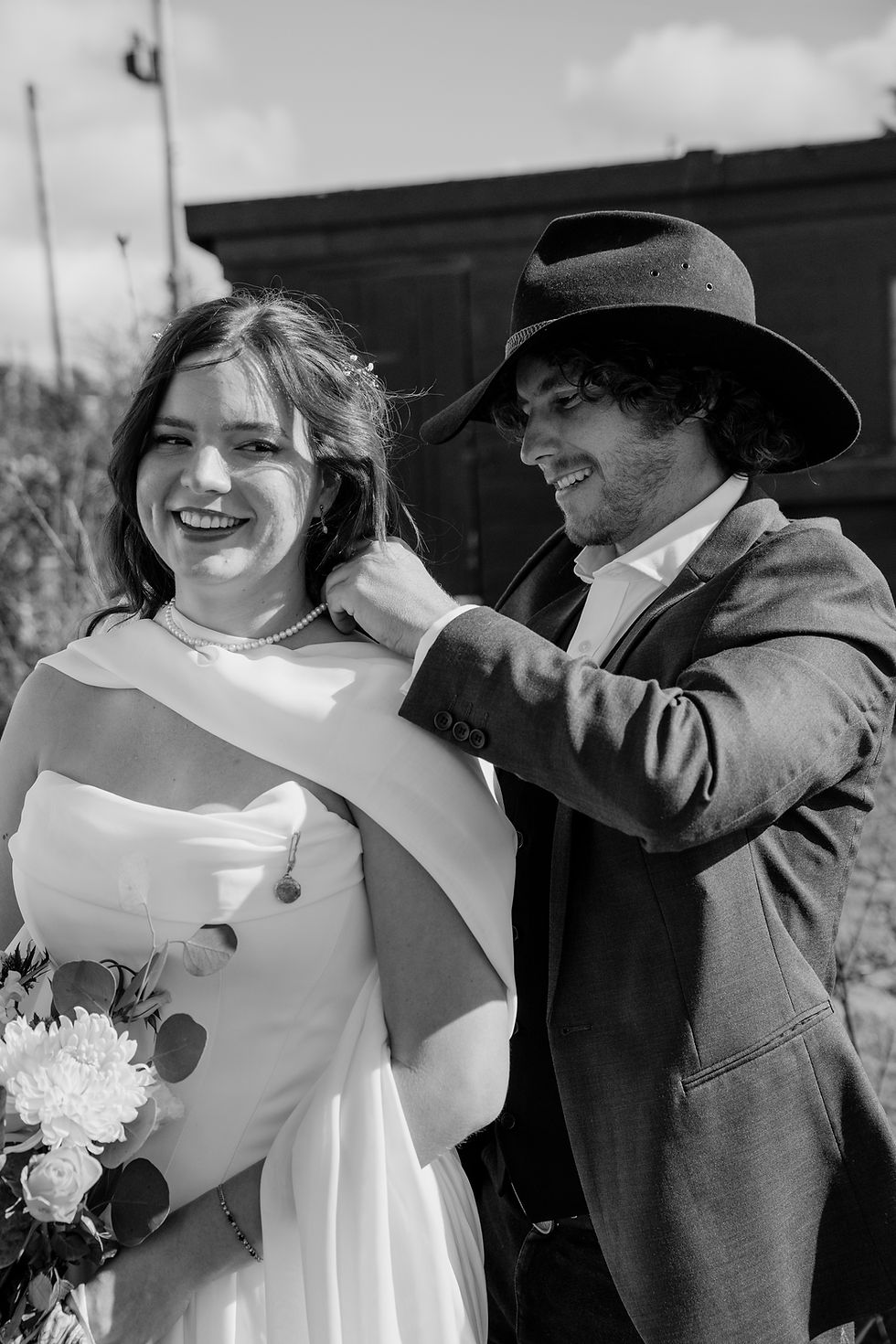 Black-and-white wedding portrait of a smiling couple outdoors. The groom places a pearl necklace on his bride, as she holds a bouquet.