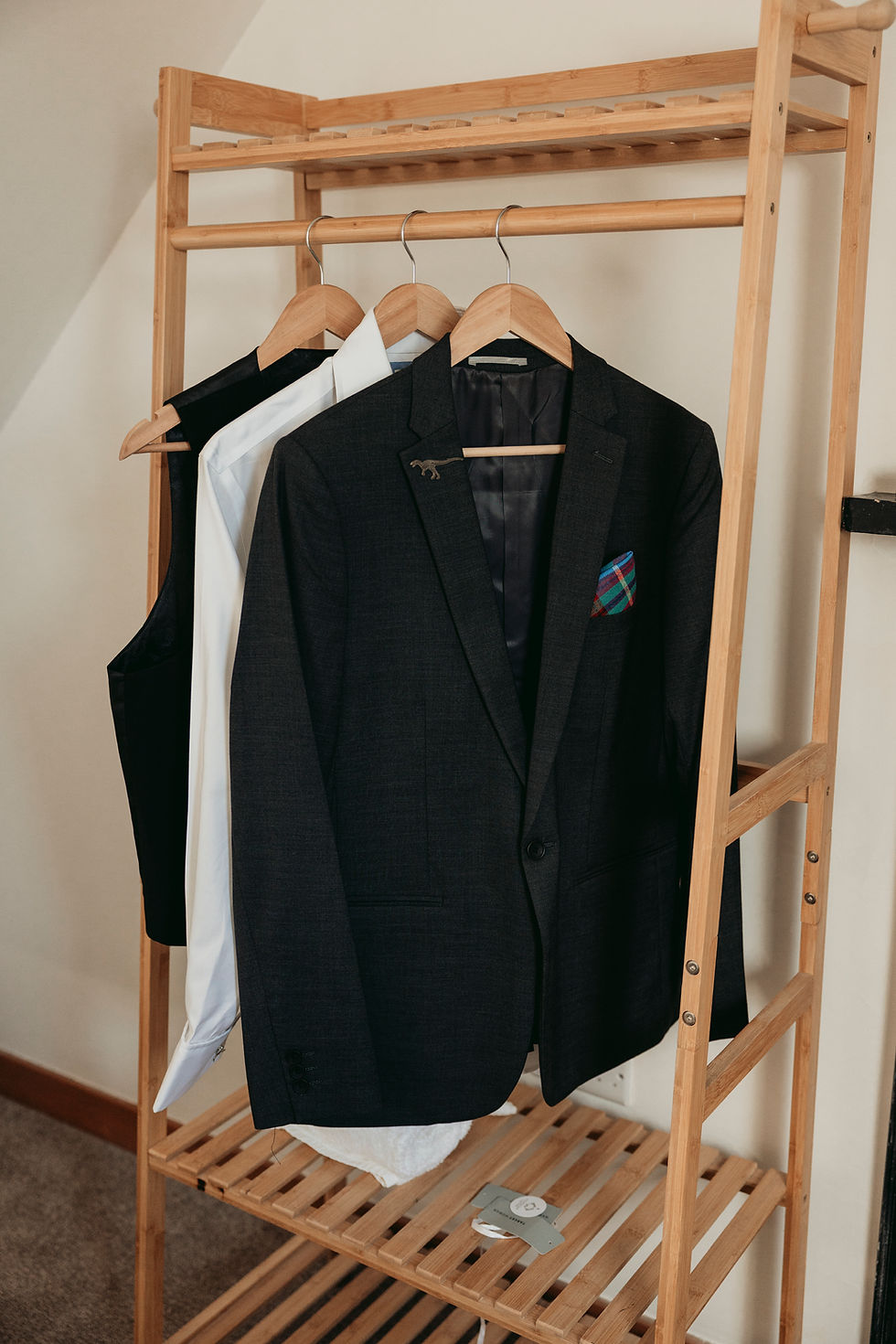 Wooden garment rack with black suit jacket, white shirt, and black vest on hangers in a tidy room.