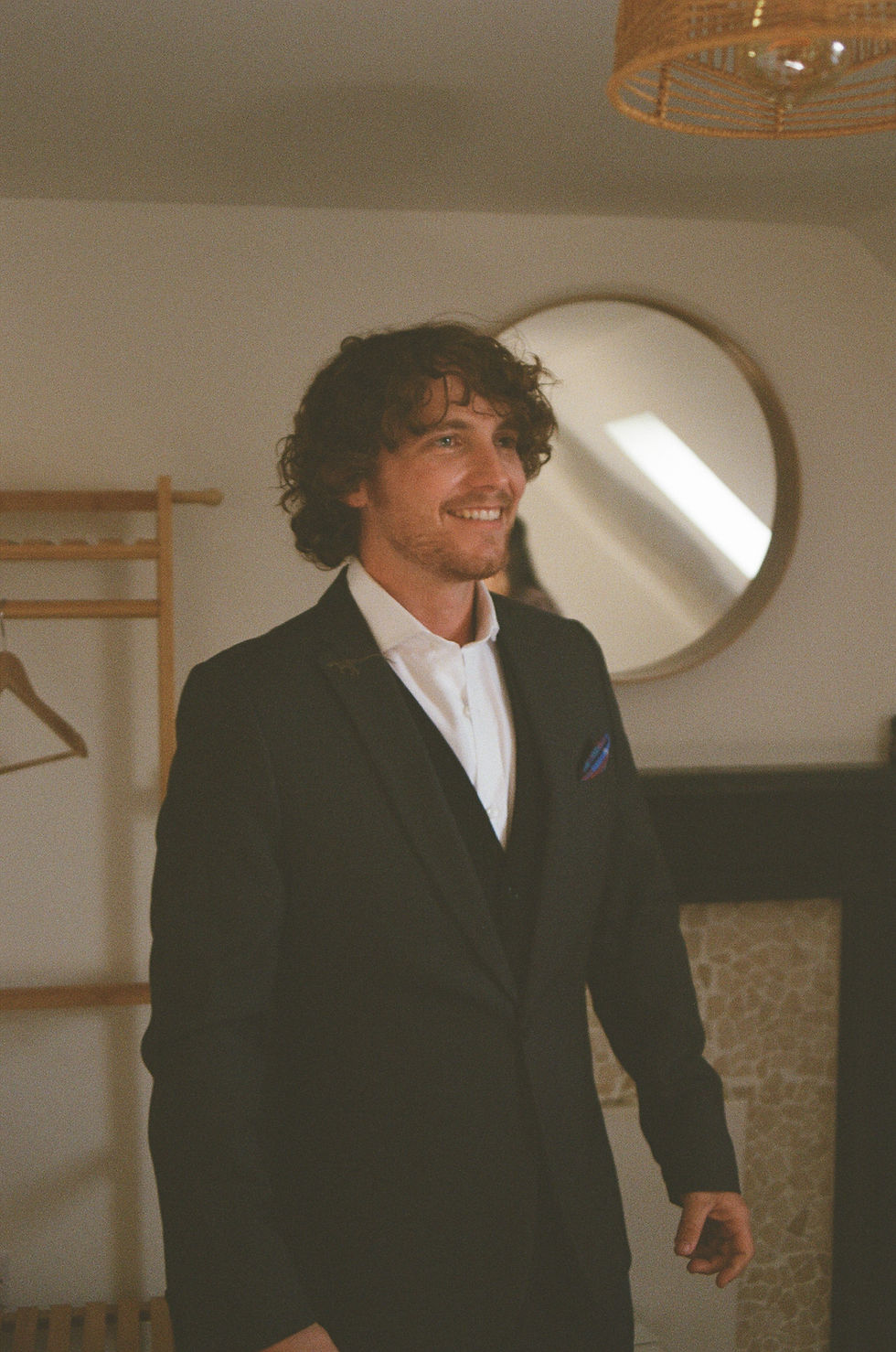 A film photo of a smiling groom in black suit and white shirt stands in a warm-lit room with a round mirror and wooden shelves.