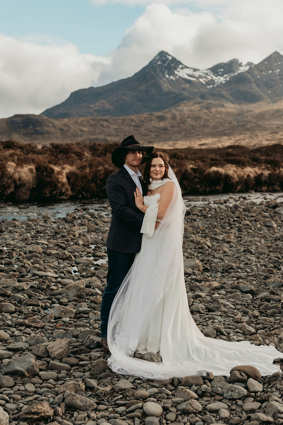 Bride and groom embrace on a rocky riverbed, the bride in a flowing white gown and veil, with mountains and clouds behind.