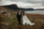 Bride and groom holding hands while walking towards the camera. Behind them are tall cliffs near Neist Point on the Isle of Skye in Scotland, as well as the ocean.