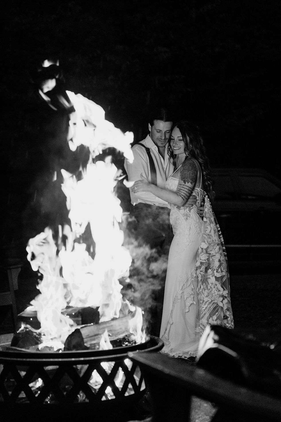 A couple embraces by a large campfire at night. The woman wears a detailed lace dress. The scene is warm and intimate.