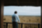 Groom in a blue shirt stands on a wooden balcony, overlooking a vast savannah of the Serengeti under a cloudy sky, evoking a sense of tranquility.