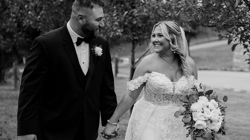 Bride and groom hold hands, smiling at each other. She's in a lace gown with a bouquet, he's in a black suit. Trees in the background.