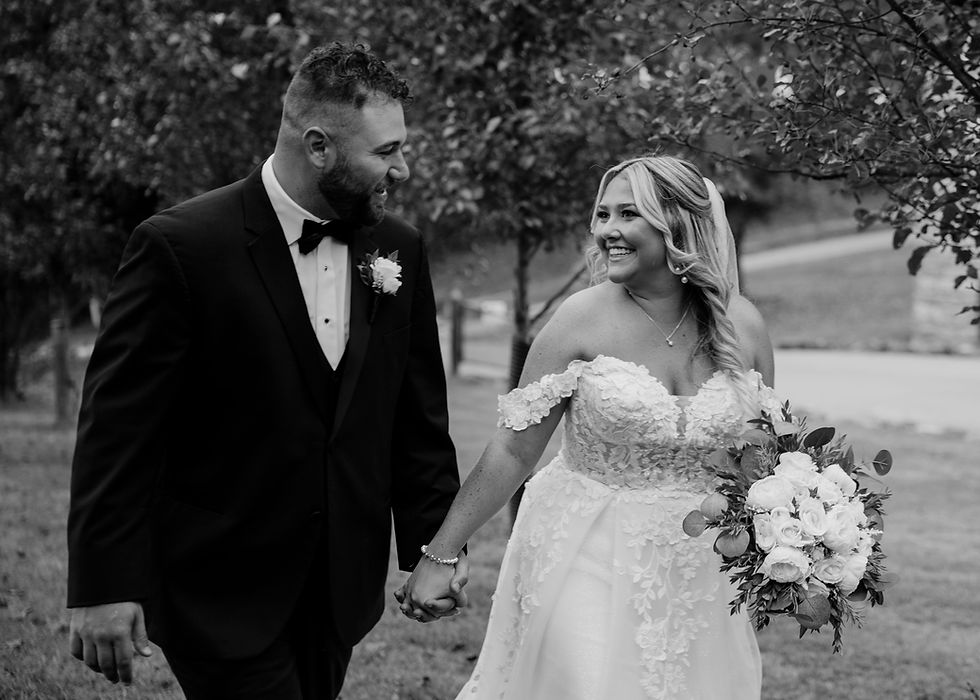 Bride and groom hold hands, smiling at each other. She's in a lace gown with a bouquet, he's in a black suit. Trees in the background.