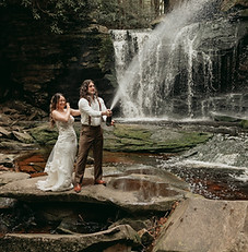 A groom pops a bottle of champagne with his bride looking excitedly behind him. They're sourrounded by mossy rocks and a tall waterfall is in the background.