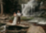 A couple joyfully sprays champagne near a cascading waterfall, standing on rocks. The bride is in a white dress, and the groom wears suspenders.