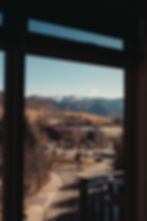 View from window of a town with brown buildings, snowy mountains in the background, clear sky, and winding road. Calm and serene scene.