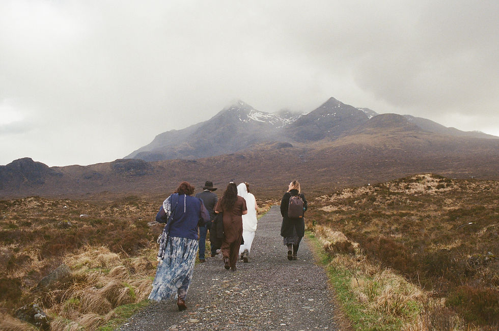 A film photo of a group of wedding guests, bride, and groom walks a gravel trail through brown moorland toward misty, snow-dusted Black Cuillin mountains of the Isle of Skye in Scotland. The sky is overcast.