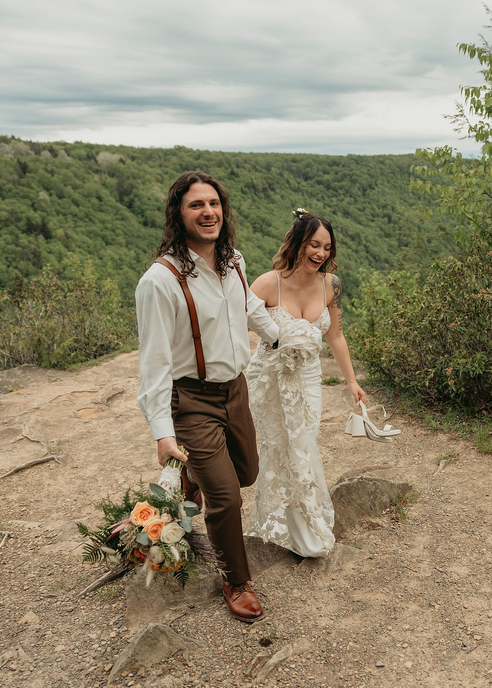 A smiling couple walks outdoors; the man holds a bouquet, and the woman carries shoes. They're on a rocky path with a lush green background.