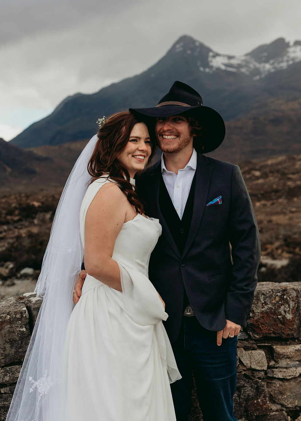 Smiling bride and groom pose on the Old Sligachan Bridge with mountains behind them; groom wears a black cowboy hat.