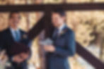 Two men in suits smiling, holding notebooks, against a rustic outdoor backdrop with trees. One book has "vows" on it. Festive mood.