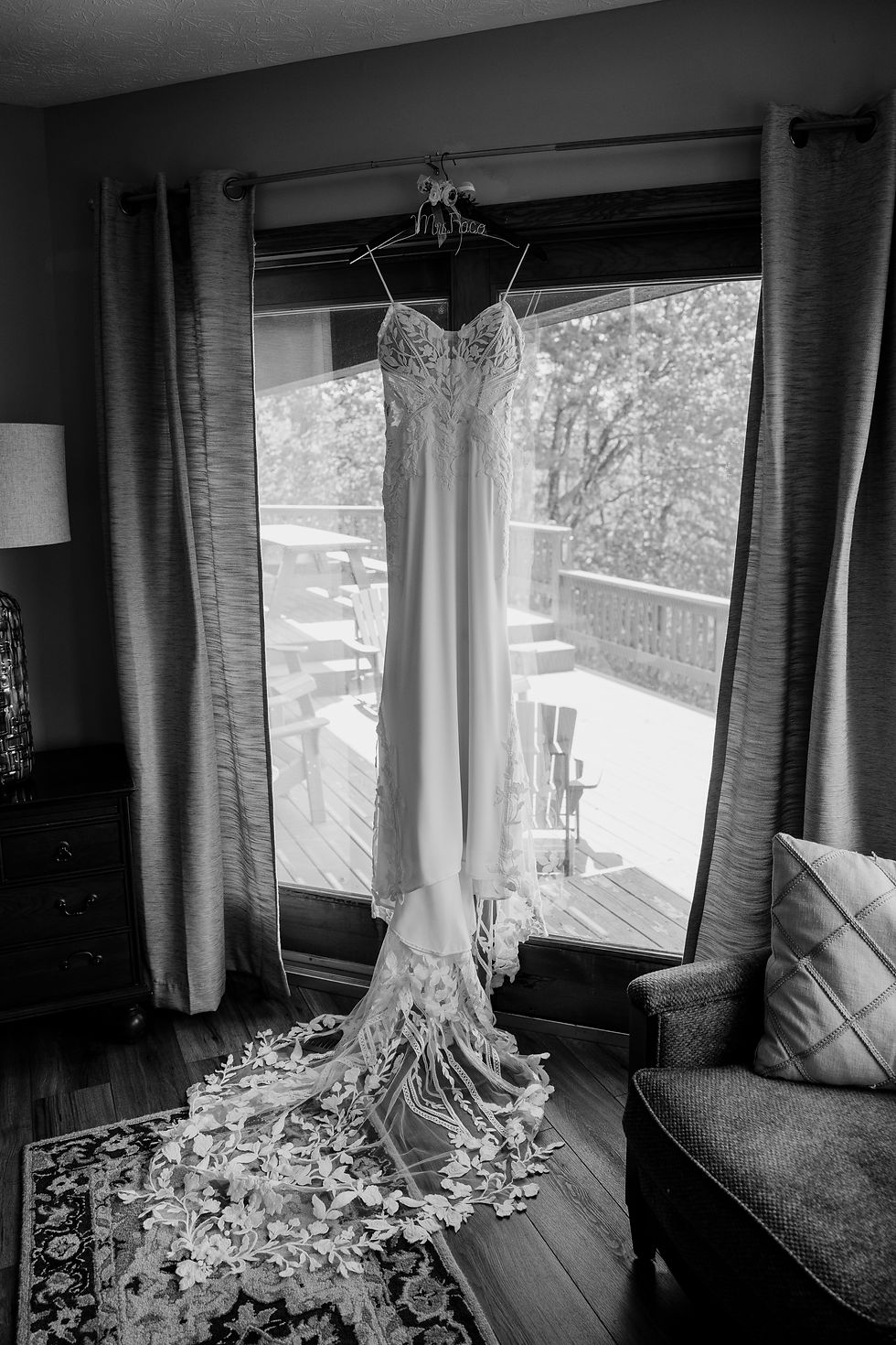 Wedding dress with lace details hangs by patio doors, overlooking wooden deck. Elegant and serene setting, monochrome tones.