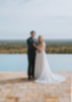 A wedding couple stands by an infinity pool overlooking a vast savanna. The bride wears a lace gown, and the groom is in a suit and pink tie.
