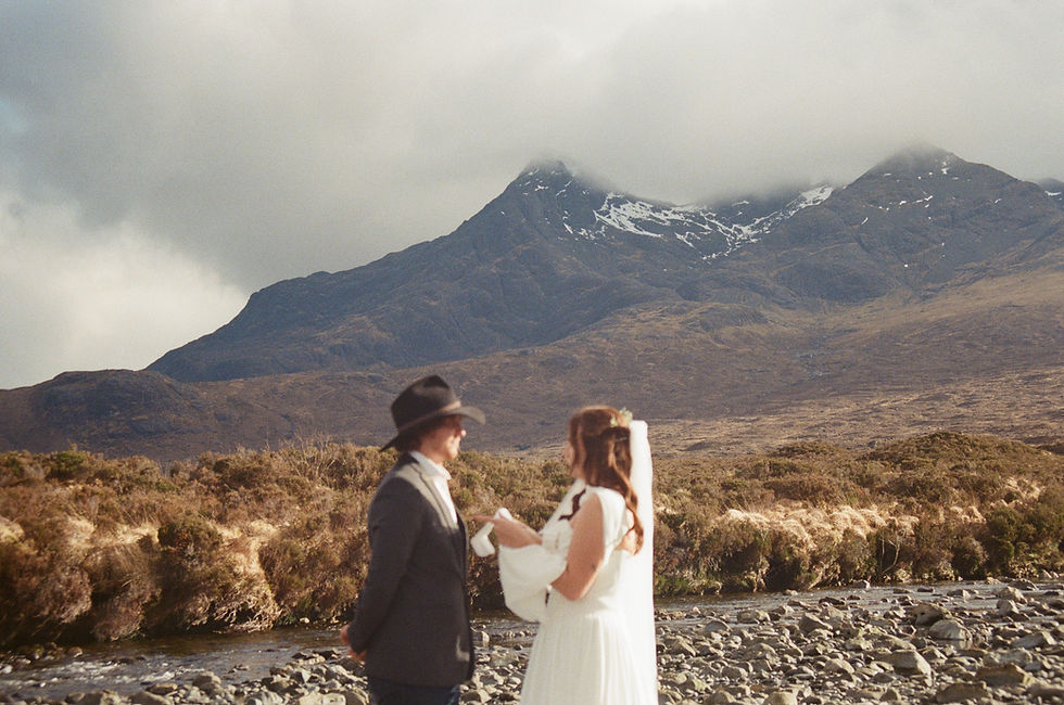 Bride and groom stand by a rocky river with misty mountains behind them, sharing a quiet wedding moment.